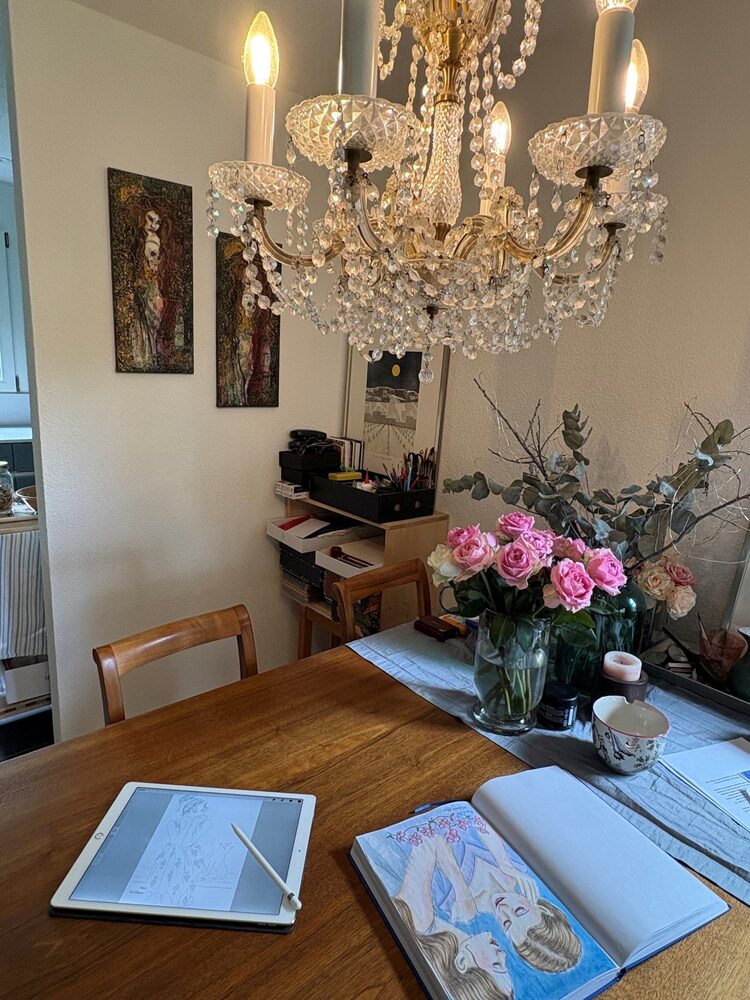 Carolyn's dining table with an iPad showing lineart, an open sketchbook with a colored drawing, a vase of pink roses and a crystal chandelier above